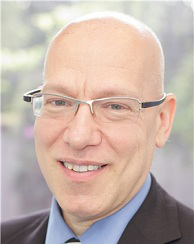 This is a photograph of a middle-aged man with glasses, wearing a dark suit and tie, smiling slightly at the camera.