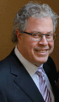 The image shows a man with glasses smiling at the camera, wearing a suit and standing against a neutral background.