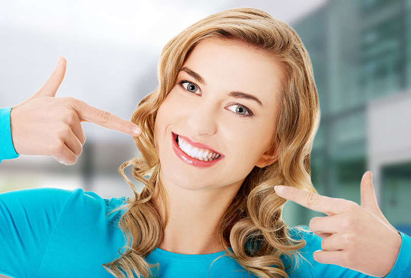 The image features a woman with blonde hair smiling broadly while pointing at her teeth with both hands. She has light skin, wears a blue top, and stands against a blurred background that suggests an indoor setting.