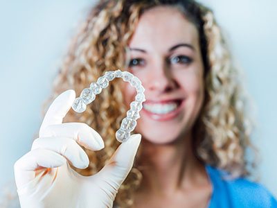 A woman holding up a clear plastic dental retainer with her left hand while wearing a white glove and smiling at the camera.