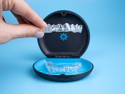 The image shows a person s hand holding an open box containing a set of clear, transparent orthodontic aligners, which are used for teeth straightening.