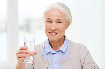 An elderly woman holding a glass of water while smiling at the camera.