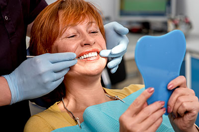 A woman with red hair sitting in a dental chair, smiling at the camera, with a dental hygienist performing a cleaning procedure on her teeth.