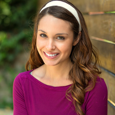 A young woman with long brown hair and a smile, wearing a purple top and a white headband, posing for a portrait outdoors.