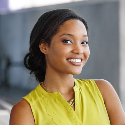 The image shows a woman with a smile, wearing a yellow top and standing against a wall, posing for a portrait.