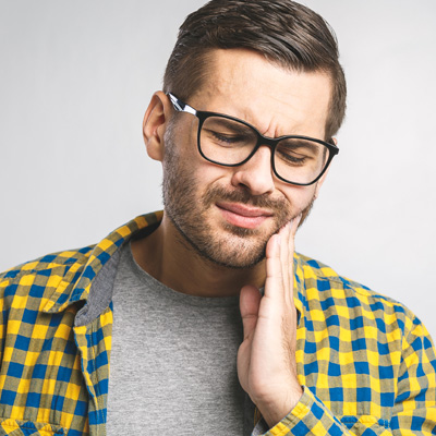 The image shows a man with glasses, wearing a yellow plaid shirt, holding his hand to his chin, and looking upwards with a concerned or questioning expression.