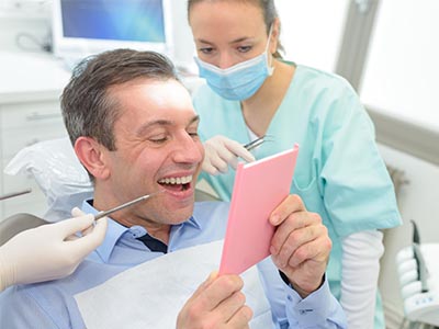 A man sitting in a dental chair with a surprised expression while holding a pink card, surrounded by dental professionals who are looking at the card with interest.