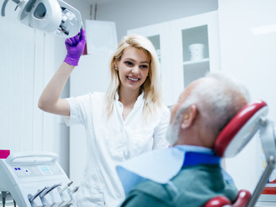 A woman in a white lab coat is standing next to a man seated in a dental chair, holding up a device with a smile on her face, both in a dental office setting.