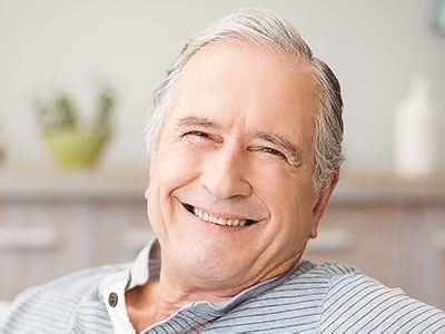 The image shows an elderly man with gray hair and a mustache, smiling at the camera, wearing a blue shirt, sitting comfortably in a chair indoors with a blurred background that suggests a home environment.