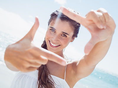 The image features a woman with her hand up to her face, holding a camera, smiling at the camera while standing outdoors on a sunny day.